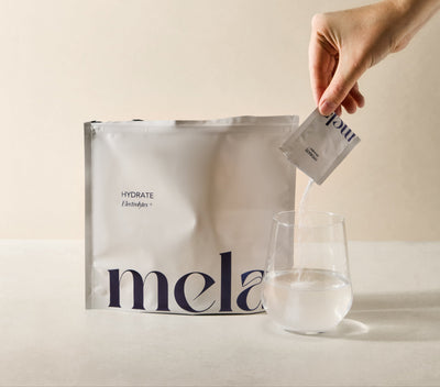 Hand pouring a packet of 'mela hydrate' into a glass with a branded bag in the background.
